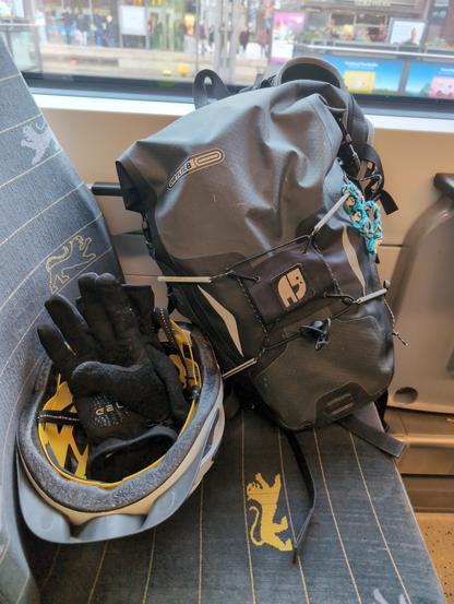 Ein Fahrradhelm mit Handschuhen und ein Fahrradrucksack liegen auf einem Sitz im Zug. Am Rucksack ist ein Patch angebracht, auf dem der Natenom-Elefant mit Träne zu sehen ist.