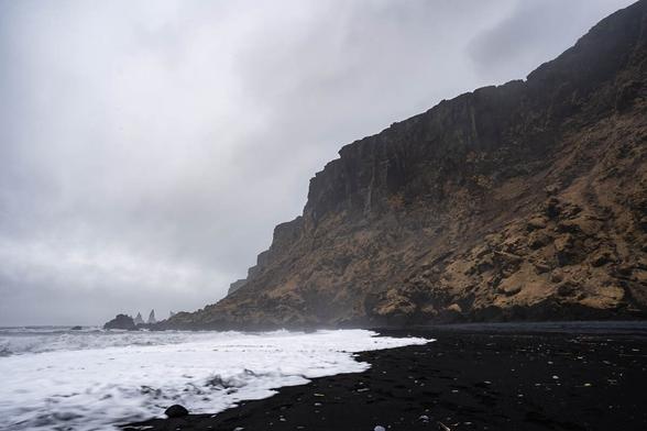 A moody scene at Reynisfjara Beach in Iceland, featuring black sand, foamy waves crashing onto the shore, and towering basalt cliffs. In the distance, the Reynisdrangar sea stacks rise from the ocean under a stormy, overcast sky.