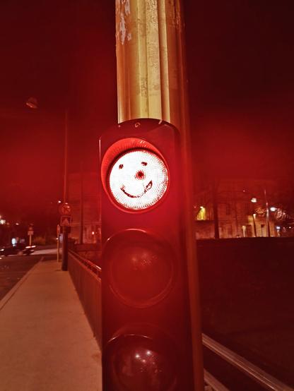 La imagen muestra un semáforo en rojo durante la noche, con una peculiaridad que lo hace destacar: en la luz roja se ha dibujado una carita sonriente 🙂. El semáforo está ubicado en una acera con barandillas a su lado, mientras que al fondo se pueden ver algunas luces de edificios y la calle parcialmente iluminada.
La escena tiene un tono cálido y un aire casi surrealista, con la luz roja proyectando un brillo intenso y reflejando sobre el poste dorado. La carita sonriente en el semáforo rompe con la seriedad del entorno urbano, añadiendo un pequeño toque de humor o ternura en medio de la noche 🚦😊.