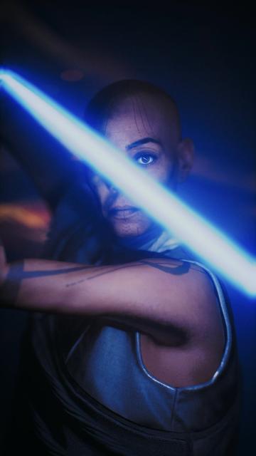 Cere with a shaved head holds a blue lightsaber, with a focused expression and visible tattoos on their face and arm. The background is dark, enhancing the glow of the lightsaber.