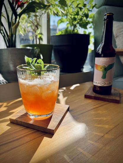 light red cocktail with mint garnish sitting on a wooden table inside with sun shining and a bottle of Braukollektiv Frida NEIPA in the background