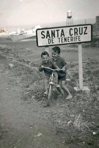 foto antigua de la entrada de santa cruz de tenerife, dos niños juegan con una bici en un camino de tierra junto a un cartel que indica la entrada a la ciudad