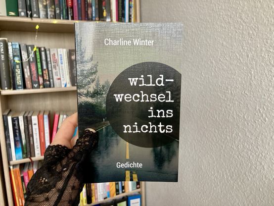 Das Buch „Wildwechsel ins Nichts“ von Charline Winter wird vor einem Bücherregal hochgehalten. Auf dem Cover ist eine verregnete Landstraße zu sehen. Der Titel steht in weißer Schreibmaschinenschrift und in Kleinbuchstaben in einem dunklen Kreis auf der rechten Seite. Ganz unten steht der Untertitel „Gedichte“.