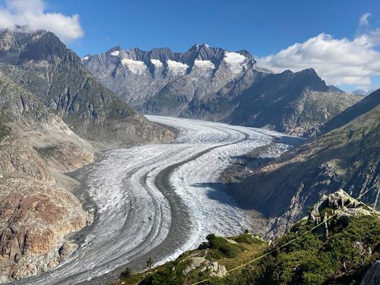 Summertime photo of the Aletsch glacier winding through a high alpine valley between rocky mountains. The glacier has alternating stripes in shades of gray and white that curve along with the flow of the glacier.
