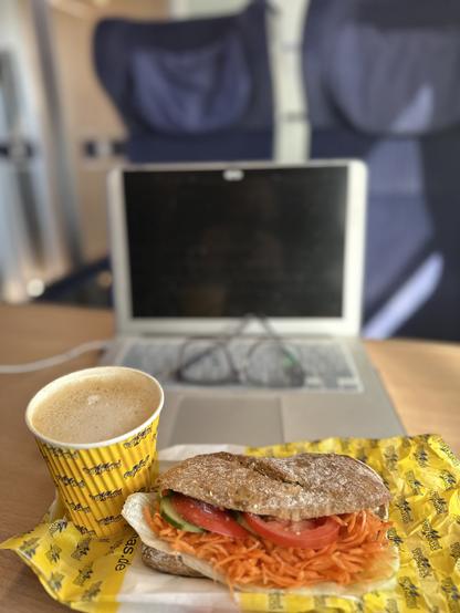 Im Vordergrund: 
Oatly Cappuccino im gelben Becher mit Haselnusssirup 2.10

Salatbaguette 3,- auf einer kleinen gelben Papiertüte. 

Beides von Yormas Essen Hbf. 

Im Hintergrund: Aufgeklappter Laptop+Brille