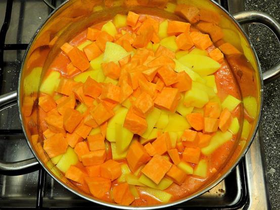 In einem großen Edelstahltopf auf einem Gasherd befinden sich gewürfelte Süßkartoffeln und Kartoffeln in einer orangefarbenen Sauce. Die Süßkartoffelwürfel sind leuchtend orange, während die Kartoffeln gelblich sind. Die Sauce hat eine cremige Konsistenz und bedeckt teilweise die Zutaten.