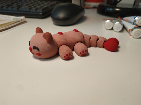 Un gattino snodabile color tortora stampato in 3d, alla fine della coda c'è un grosso cuore rosso e due cuoricini invece sono sulla schiena. Faccino sorridente con nasino a cuore e baffetti marroncini come le orecchie.