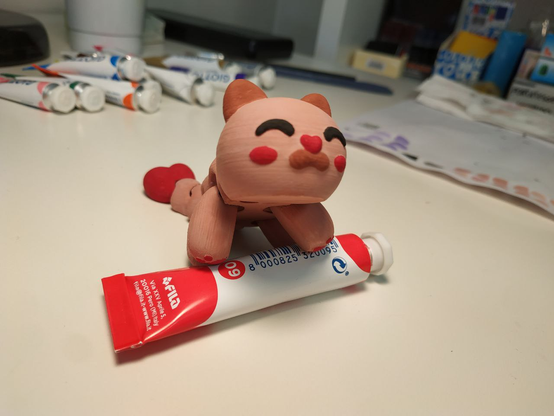 Stesso gattino appoggiato con le zampine su un tubetto di tempera rossa, un po' di bordello sullo sfondo.