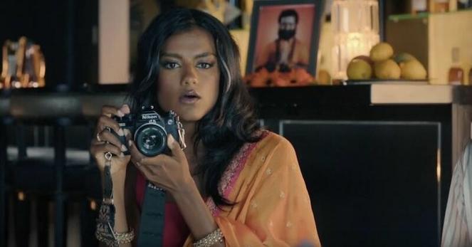 Simone Ashley holds a camera and is looking at her subject. Behind her are lots of photos and a bowl of oranges.