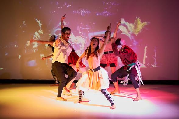 The photo shows a group of 6 dancers on stage dancing wildly. The background and the whole scene is illuminated very colorful in orange, yellow, red and pink.