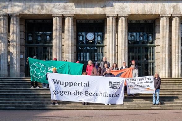 Rund 20 Menschen stehen tagsüber vor einem historisch anmutenden Gebäude mit hohen Türen und Säulen-Fassade. Sie halten Banner mit der Aufschrift „Wuppertal gegen die Bezahlkarte“ und „Menschenwürde ist migrationspolitisch nicht zu relativieren – BverfG, 18.07.2012“
