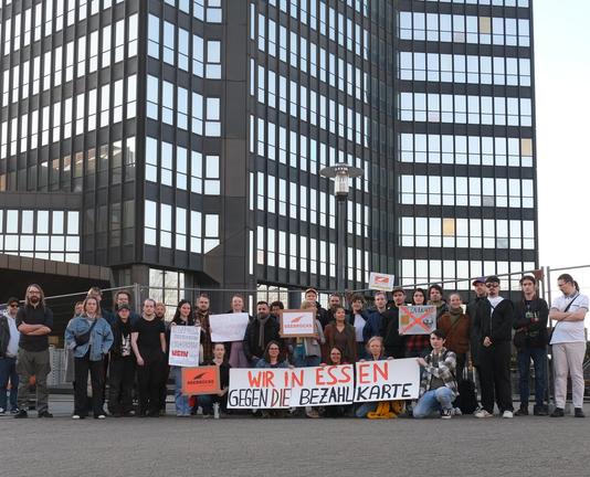 Rund 30 Personen stehen vor einer Hochhausfassade, dunkel mit hell verspiegelten Fenstern. Sie halten ein großes Pappschild mit der Aufschrift „Wir in Essen gegen die Bezahlkarte“