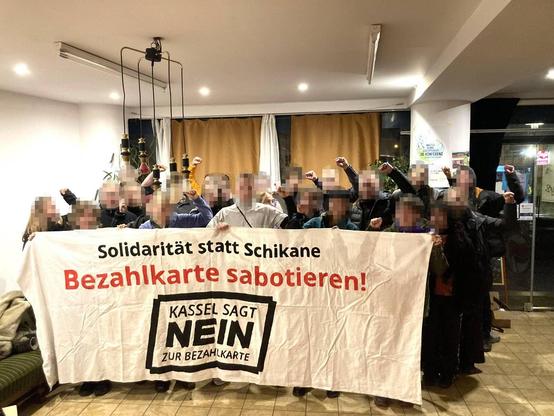 Rund 20 Menschen stehen in einem hell erleuchteten Innenraum hinter einem Banner mit der Aufschrift „Solidarität statt Schikane – Bezahlkarte sabotieren! Kassel sagt Nein zur Bezahlkarte“. Ihre Gesichter sind verpixelt, doch man erkennt ihre kämpferisch erhobenen Fäuste.