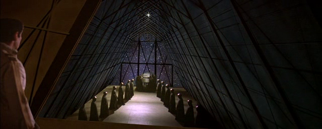 La imagen muestra el interior de una estructura piramidal con paredes oscuras y un techo de cristal que simula un cielo nocturno estrellado. En el centro, se observa un ataúd o una urna sobre una plataforma elevada, rodeado de personas vestidas con túnicas oscuras que parecen estar de luto o en una ceremonia fúnebre. Una persona, de espaldas a la cámara, se encuentra en la esquina superior izquierda observando la escena. La atmósfera es solemne y oscura.