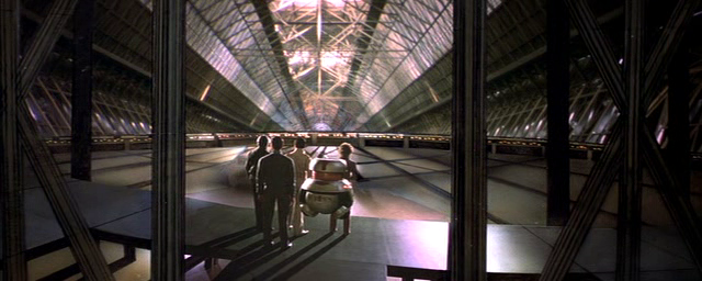 La imagen muestra un interior futurista y amplio, parecido a una galería o atrio, con una estructura de techo de cristal y acero que deja pasar mucha luz.  Cuatro figuras humanas, vistas de espaldas, están de pie frente a un pequeño droide o robot cilíndrico.  Las figuras parecen estar observando el droide. El espacio está definido por una estructura de metal oscuro y brillante que crea un efecto de pasillo o pasarela. La perspectiva es desde una posición ligeramente elevada, observando la escena desde un punto de vista lateral.