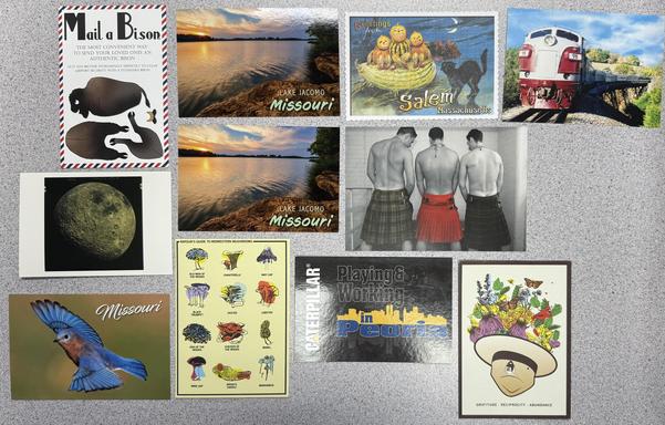 A number of postcards arranged on a table. Mail a bison, a train, a photo of the dark side of the moon, a Missouri bluebird amongst others.