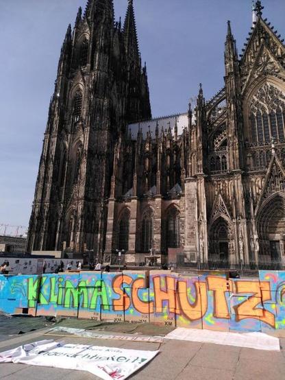 Das Bild zeigt die Kölner Dom, ein imposantes gotisches Bauwerk mit seinen charakteristischen spitzen Türmen und kunstvollen Fassaden. Vor dem Dom befindet sich eine provisorisches Fundament aus Kartons, auf der in bunten Farben die Aufschrift "Klimaschutz" in großen Buchstaben gemalt ist. Die Farben sind lebhaft und variieren von Grün, Orange bis Blau. Darunter liegen weitere Plakate und Stoffe. Im Hintergrund sind einige Menschen zu sehen, die sich in der Nähe des Doms aufhalten. Der Himmel ist klar und blau.