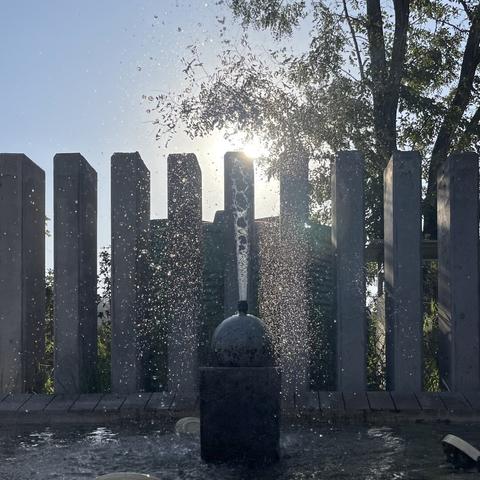 Square photo of a fountain shooting water up, with 9 concrete pillars and a tree being all backlit by the late afternoon sun. The water flow starts breaking up just a few centimeters from the ejection point.