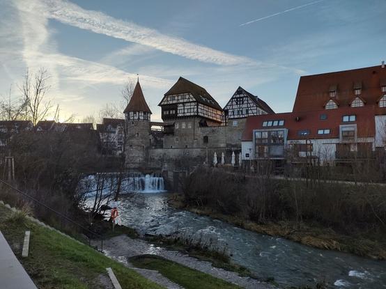Das Bild zeigt eine historische Altstadt mit beeindruckenden Fachwerkhäusern, die auf einer alten Steinmauer errichtet wurden. Im Vordergrund fließt ein Fluss mit einem kleinen Wasserfall, der über eine Staustufe hinabstürzt. Links neben dem Wasserfall steht ein runder, mittelalterlicher Turm mit einem spitzen Dach, der Teil der alten Stadtbefestigung zu sein scheint.
Rechts des Turms erstreckt sich ein großes, markantes Fachwerkgebäude mit einem steilen Dach, das auf einem massiven Steinunterbau ruht und teilweise über die Mauer hinausragt. Weitere Fachwerkhäuser mit roten Ziegeldächern und großen Fenstern befinden sich daneben, darunter ein moderneres Gebäude mit großen Glasflächen.
Der Himmel ist leicht bewölkt mit langen, weißen Wolkenstreifen, und die tief stehende Sonne wirft ein warmes, goldenes Licht auf die Gebäude, während die Bäume im Vordergrund kahl sind.