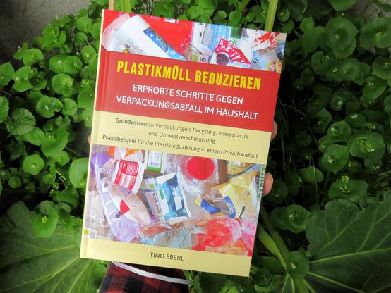 Eine Hand hält ein Buch mit einem Cover, das Bilder von Plastikverpackungen zeigt. Der obere Teil des Buchumschlags ist rot und der untere beige. Im Hintergrund befinden sich Rhabarberpflanzen und Winterportulak in einem Kübel in dichter Anordnung.