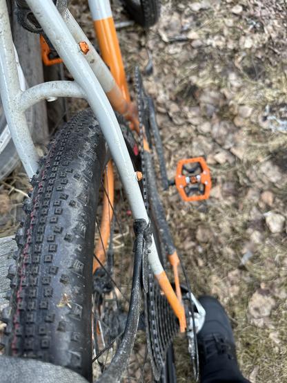 Harmaaoranssi pyörä, jossa paksu kuvioitu takarengas, oranssi hybridipoljin ja tumma pyöräilykenkä pyörän vieressä näkyvissä. Kuva on rajattu niin, että pyörästä näkyy takapyörä ja polkimet takaa ylhäältä kuvattuna