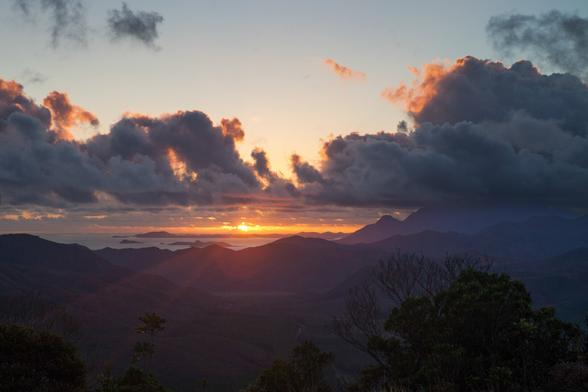 A stunning sunset over a mountainous landscape, with vibrant colors in the sky and silhouetted peaks extending into the horizon, partially obscured by clouds.