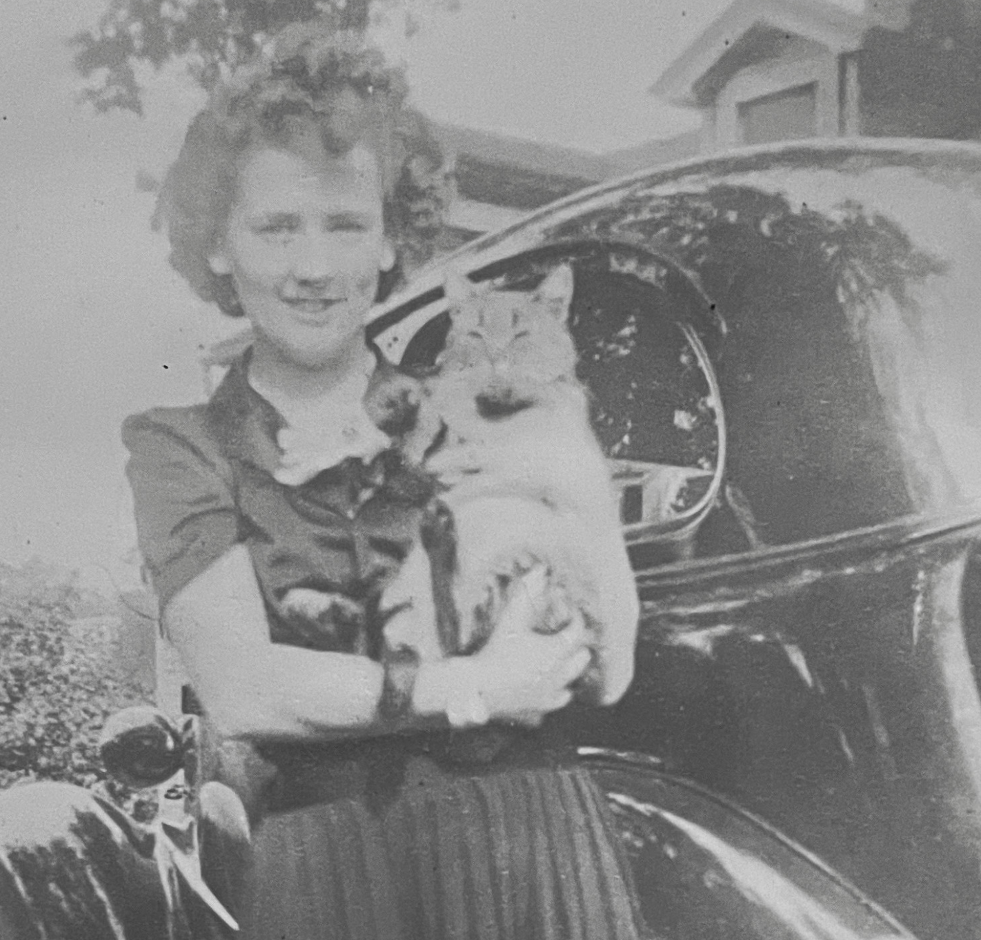 Photo of a young light skinned woman with curly hair, cradling a cat in her arms on its back like a baby. They are standing outside in front of a car.