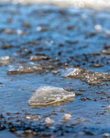 The image shows pieces of ice on a wet, possibly muddy surface. The focus is primarily on a large piece of ice in the foreground, with more ice and wet ground visible in the background. The setting appears to be outdoors, possibly in a place where ice is melting.