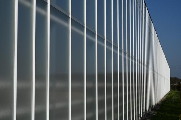 ein Gewächshaus, bestehend aus vielen Reihen von (Plexi)Glasscheiben. Die Kamera wurde relativ dicht an das Motiv geführt, sodass die senkrechten Verbindungsstege (Alu) zwischen den Scheiben nach hinten immer dichter zusammen rücken. So entsteht ein netter geometrischer Effekt, der Spannung ins Bild bringt. Die Sonne durfte von hinten scheinen, für ein bisschen Bling, und der Himmel war schön blau