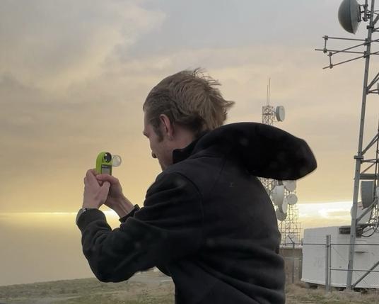 A man facing away from the camera struggling at stand in strong wind while holding a handheld anemometer. Radio towers are visible behind him.