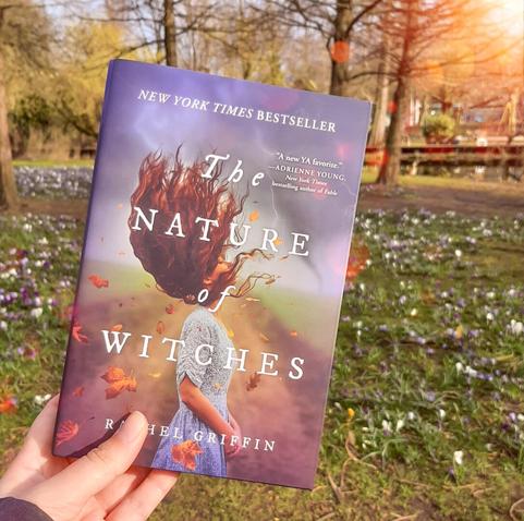 Hand holding the book The Nature of Witches up outside in front of a meadow with spring flowers