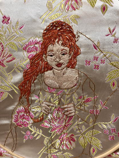 An embroidered portrait of a pale skinned red haired woman looking down toward her hands.