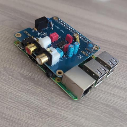 Una placa de circuito impreso azul, probablemente una tarjeta de sonido o similar, se encuentra sobre una superficie de madera gris. La placa tiene varios componentes electrónicos, incluyendo condensadores, resistencias y conectores RCA dorados. También se ven dos puertos USB y un puerto Ethernet. La placa está etiquetada como "Pi HAT" y "RPi DAC+".