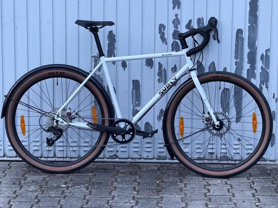 A white Surly bike is positioned against a weathered metal wall. It features thick tires with brown accents, a sleek frame, and standard bike components, including brakes and a gear system.
