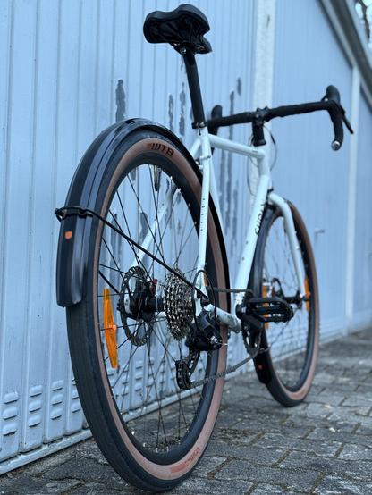 A close-up view of a bicycle, showcasing its rear angle. The bike features a white frame, black handlebars, and distinctive tires with tan sidewalls. The background consists of a textured gray wall.