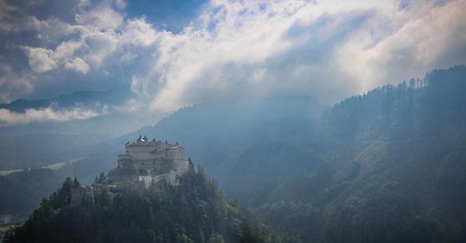The image shows a majestic castle set atop a hill surrounded by lush green forests. In the background, there are mountains partially obscured by clouds and mist, creating a dramatic and ethereal atmosphere. The sunlight piercing through the clouds adds a mystical quality to the scene.