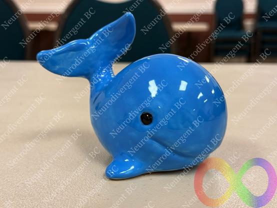 A small, glossy blue ceramic whale figurine is placed on a table. It has a rounded body and a distinct tail. The background includes blurred green chairs. Watermarks of "Neurodivergent BC" and a colourful infinity symbol are over it.