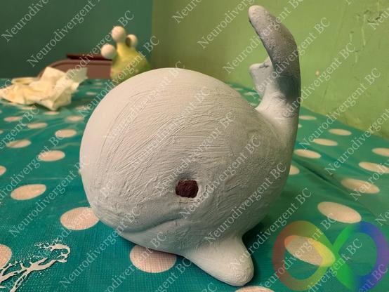 These photos show a hand-painted ceramic or clay whale figurine, possibly in the process of being painted. The whale is painted in a light pastel blue shade with some visible brush strokes and has small, round, dark eyes. The setting looks like a crafting or art space, with a polka-dotted tablecloth and other painted objects in the background.