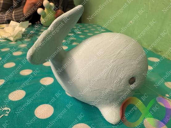 A light blue, unfinished ceramic whale figurine sits on a polka-dotted tablecloth. In the background, a person holds a green figure, and the setting features a green wall.