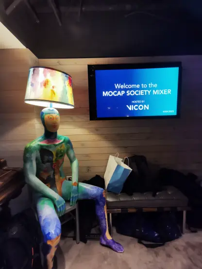 A corner of the event space featuring a life-sized mannequin painted in bright colors and abstract patterns. The mannequin is seated on a chair with its arms resting on its legs. A lamp with a colorful lampshade is attached to its head, illuminating the area. Behind it, a screen displays the "MOCAP SOCIETY MIXER" welcome message. Several bags are placed on the bench next to it.