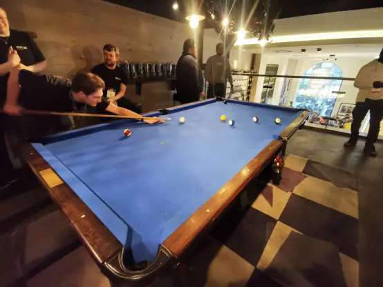 A billiards table in an elevated section of the venue, surrounded by several people. A man is in the middle of taking a shot with a pool cue, while others watch. In the background, attendees are socializing, and the space is well-lit compared to the lower level of the venue.