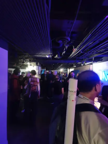 A crowded indoor networking event with attendees standing in small groups, engaged in conversation. Many are wearing badges around their necks, indicating their participation in the event. The space is dimly lit, with purple-tinted lighting. The ceiling has exposed pipes and ventilation ducts.