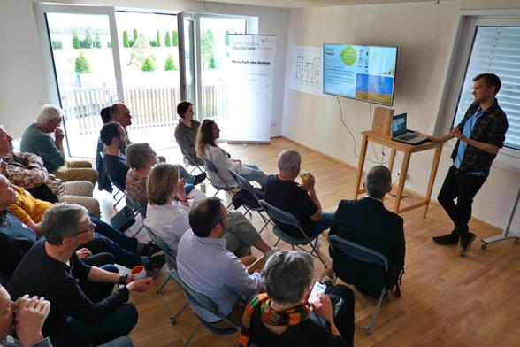 Ein Raum mit einigen Leuten. Vorne steht ein Referent mit Laptop am Stehpult; der Bildschirm mit seiner Präsentation ist ebenfalls zu sehen.
