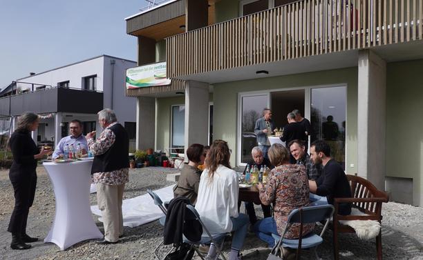 Vor einem Haus sitzen einige Personen ins Gespräch vertieft um einen runden Tisch in der Sonne, daneben ein Stehtisch mit drei Personen im Gespräch