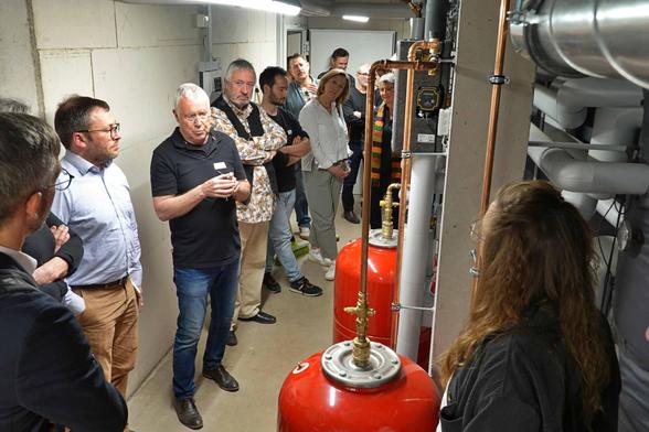 Einige Menschen des Gemeinwohlökonomietreffens im Heizungskeller mit Rohren und den großen Heißwassertanks, in denen der durch Solarthermie erwärmte Heißwasservorrat ist. Im Vordergrund Gunnar Laufer-Stark, Vorstand der nestbau AG.