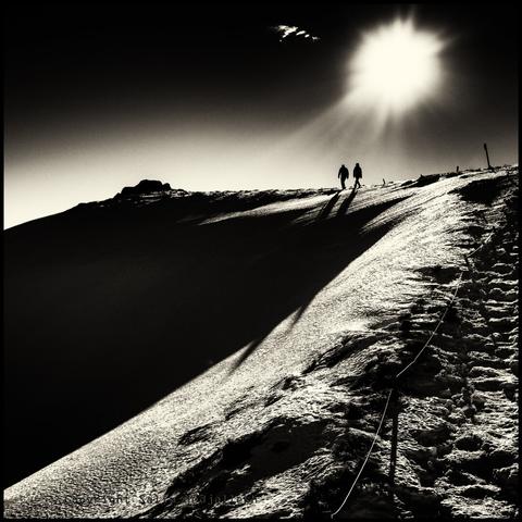 Photographie noir et blanc ascension du Puy de Sancy sous la neige en contre jour avec 2 personnes sur une crête.
Black and white photograph of a snow-covered ascent of Puy de Sancy with 2 people on a ridge.