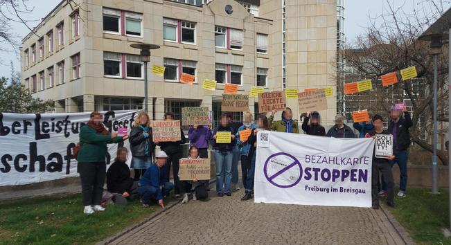 Viele Menschen stehen draußen vor Haus.
Sie haben zwei Banner und viele Papp- und bunte Papierschilder, die sich gegen die Bezahlkarte und das Asylbewerberleistungsgetz richten.
Auf dem linken Banner steht: Asylbewerberleistungsgesetz abschaffen.
Auf dem vorderen Banner steht: Bezahlkarte stoppen Freiburg im Breisgau, daneben ist eine durchgestrichene Bezahlkarte abgebildet.
Auf den Schildern stehen u.a.:
- Landratsamt, ihr seid die Vollstrecker rassistischer Politik
- Verantwortung beginnt regional
- Gleiche Rechte für alle!
- Ausgrenzung ist kein Versehen, sie ist System
Nicht alle Schilder sind lesbar.