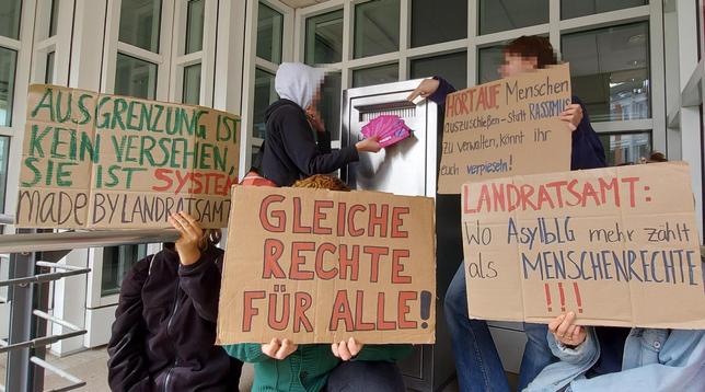 Mehrere Menschen halten Schilder hoch vor dem Landratsamt. Eine Person wirft Flyer mit "Nein zur Bezahlkarte" in den dortigen Briefkasten.
Auf den Schildern steht:
. Ausgrenzung ist kein Versehen, sie ist System made by Landratsamt
- Gleiche Rechte für alle!
- Landratsamt: Wo AsylbLG mehr zählt als Menschrechte!!!
- Hört auf Menschen auszuschließen, statt Rassismus zu verwalten, könnt ihr euch verpieseln!