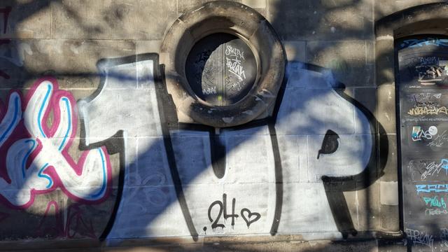 Großes Grafitti an einer Wand. "1Up" in weiß mit schwarzer Umrandung. Im U steht "24 🖤"