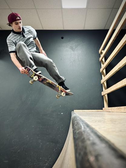 A male skateboarder in a red backwards hat, blue and gray striped polo shirt, gray pants, and black shoes performs a frontside nose grab on a larger indoor quarter pipe.